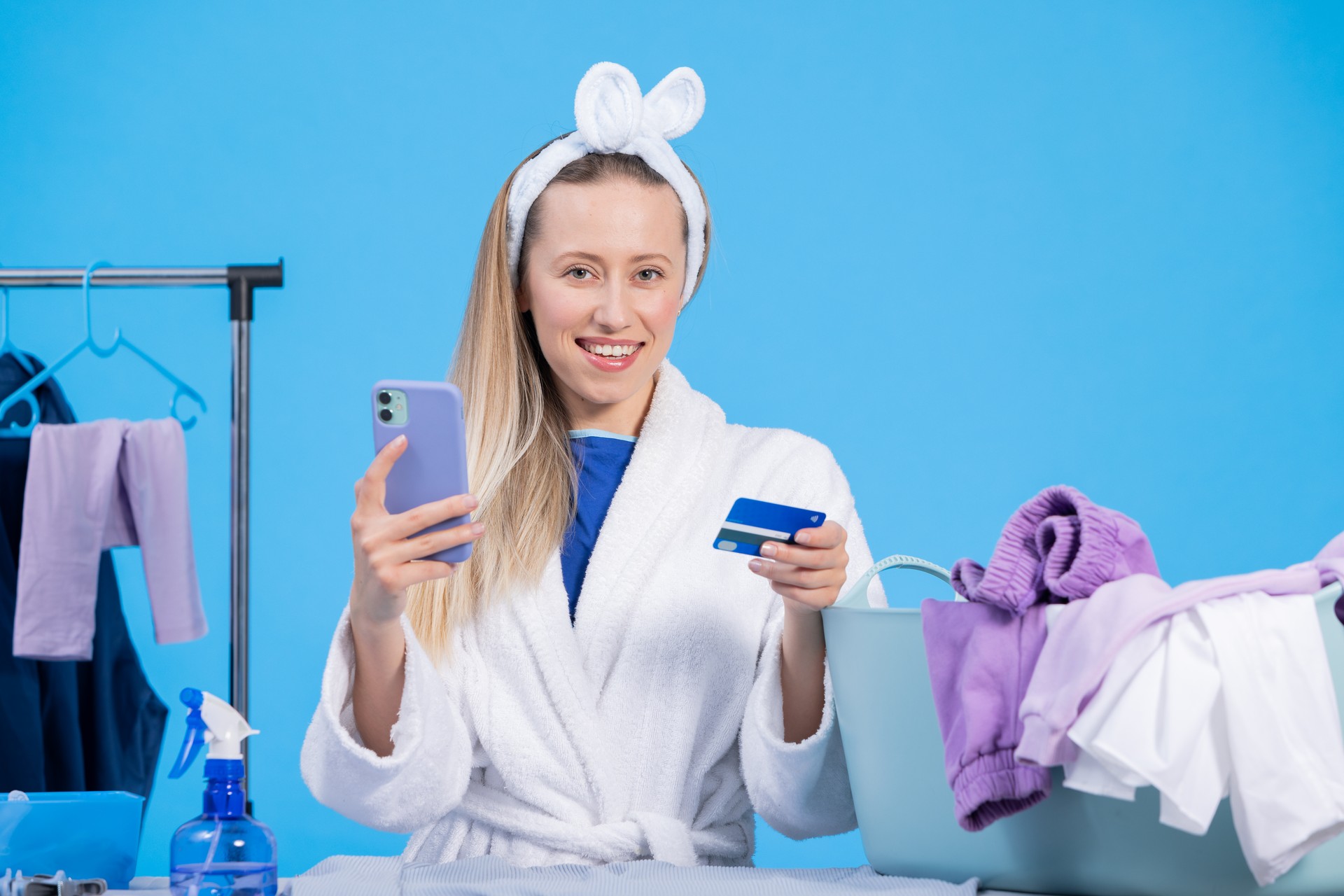 Excited funny young female housekeeper 20s in white bathrobe holds phone with credit card while doing housework isolated on pastel blue color background studio portrait. Housekeeping concept